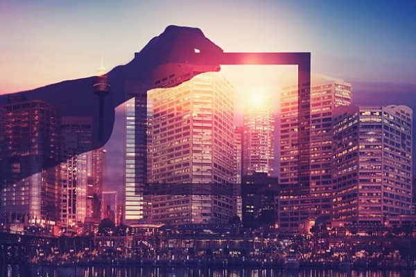 Business man holding a picture frame over city. The man is unrecognizable and you cannot see his face. He is superimposed onto a city skyline at sunset. He is holding a picture looking into the city. Success, vision, innovation concept with copy space.